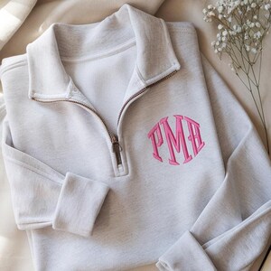 Könnte beinhalten: Ein hellgraues Sweatshirt mit einem Viertel-Reißverschluss-Ausschnitt und einem rosa Monogramm. Das Sweatshirt hat lange Ärmel mit gerippten Bündchen. Ein Zweig weißer Blumen ist neben dem Sweatshirt platziert.