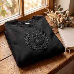 May include: Black sweatshirt featuring a cat design. The cat is holding a cup, surrounded by a floral wreath. The design is embossed on the front of the sweatshirt. The sweatshirt is folded on a wooden surface near a window.