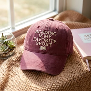 May include: A maroon baseball cap with the embroidered text "READING IS MY FAVORITE SPORT" and a small book graphic. The hat is on a tan blanket next to a pink journal and a small glass terrarium.