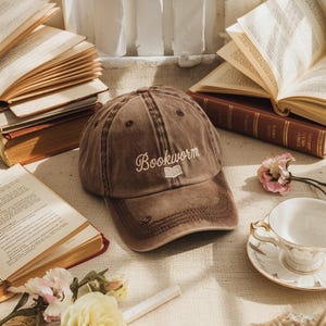 Może przedstawiać: Brązowa czapka bejsbolowa z napisem "Bookworm" wyhaftowanym białą czcionką, wraz z małą grafiką książki. Czapka jest postarzana, z zakrzywionym daszkiem i klasycznym sześciopanelowym designem. Książki i filiżanka herbaty w tle.