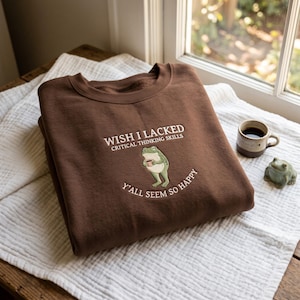 May include: A dark brown crewneck sweatshirt featuring an embroidered graphic of a green frog holding a small mug. The text above the frog reads WISH I LACKED CRITICAL THINKING SKILLS and the text below reads Y'ALL SEEM SO HAPPY in white.