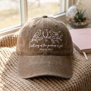 Op de afbeelding: Bruine baseballcap met een gebogen klep. De cap heeft een witte afbeelding van drie vogels op een tak, met de tekst "I will sing of the goodness of god" en "PSALM 89:1". De cap is gemaakt van een zacht, getextureerd materiaal.