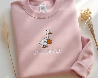 May include: Pale pink sweatshirt with a round neck and long sleeves. The design features an embroidered cartoon duck holding bread, with the text "IS THIS GLUTEN FREE?" underneath. A casual, comfortable garment.