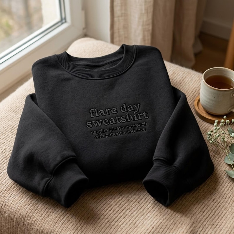 Puede incluir: Una sudadera negra de cuello redondo con bordado negro tonal en el pecho. El texto dice "flare day sweatshirt" seguido de "caution please approach with patience and love" en un tama&ntilde;o de fuente m&aacute;s peque&ntilde;o debajo.
