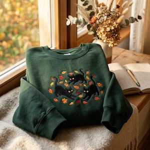 Puede incluir: Una sudadera de cuello redondo en color verde bosque oscuro con un diseño bordado de tres gatos negros jugando entre hojas de otoño dispersas en tonos naranja, amarillo y rojo. La prenda de manga larga tiene cuello y puños acanalados.