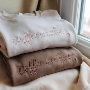 May include: Two stacked sweatshirts, one beige and one brown, embroidered with the words "coffee weather" and a heart. The sweatshirts are folded, suggesting a comfortable, casual style.