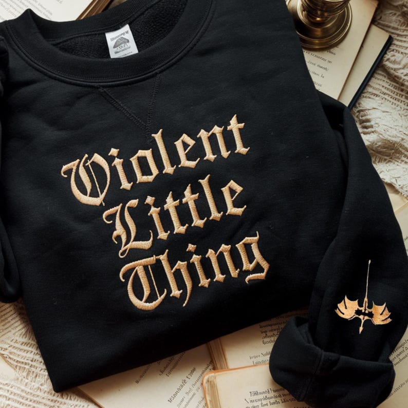 May include: Black sweatshirt with the embroidered text "Violent Little Thing" in a vintage-style font. A small gold-colored emblem is on the sleeve. The sweatshirt is displayed on top of open books.