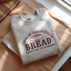 May include: Beige sweatshirt with the embroidered text "I BAKE MY OWN SOURDOUGH BREAD & DON'T TRUST THE GOVERNMENT" and a bread loaf graphic. The sweatshirt is folded on a light brown surface.
