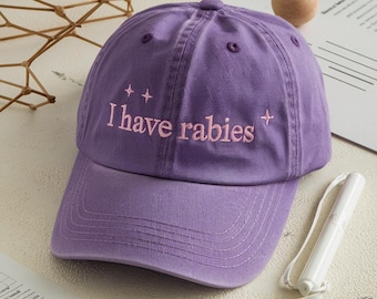 May include: A purple baseball cap with the embroidered text "I have rabies" in a lighter purple color. The cap has a curved brim and a six-panel design. The background includes a wooden sphere and a white object.