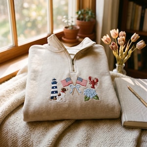 May include: A beige quarter-zip sweatshirt featuring embroidered designs on the front. The embroidery includes a blue and white lighthouse, a brown and white dog, two American flags tied with a light blue ribbon, a red lobster, and blue hydrangeas.