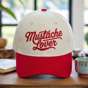 Pode incluir: Um boné de beisebol na cor creme com aba vermelha e um botão vermelho no topo. A parte frontal do boné apresenta o texto bordado "Mustache Lover" em uma fonte cursiva vermelha e ousada. O boné possui uma copa estruturada e ilhós costurados.