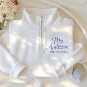 May include: White pullover sweatshirt with a quarter-zip collar. The sweatshirt features the embroidered text "Mrs. Anderson" in blue script, along with the wedding date "EST. 10.29.2025" and two wedding rings. A ring box with a diamond ring is visible.