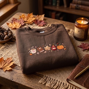 Puede incluir: Una sudadera de cuello redondo color marrón oscuro con un bordado horizontal central. El diseño muestra una taza blanca con un corazón rosa, un libro marrón, una hoja de arce, una vela roja y una calabaza naranja, con pequeñas estrellas.