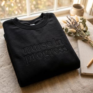 May include: A black crewneck sweatshirt featuring the text "nurse in progress" embroidered in a matching black thread across the chest. The garment has a ribbed collar and long sleeves, showcasing a clean, monochromatic design throughout.