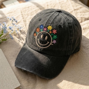 Könnte beinhalten: Eine schwarze Baseballkappe mit gebogenem Schirm. Die Kappe hat ein gesticktes Smiley-Gesicht mit einem bunten Blumenkranz. Der Hut ist aus einem weichen, abgenutzten Material gefertigt.