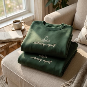 Puede incluir: Dos sudaderas de cuello redondo en color verde bosque apiladas una sobre otra. Cada sudadera presenta un gráfico bordado de un torno de alfarero con un jarrón y el texto "centering myself" en hilo de color claro centrado en el pecho.