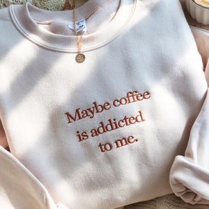 May include: A light beige sweatshirt with the embroidered phrase "Maybe coffee is addicted to me." A gold necklace with a circular pendant is draped over the neckline. The sweatshirt has a crew neck and long sleeves.