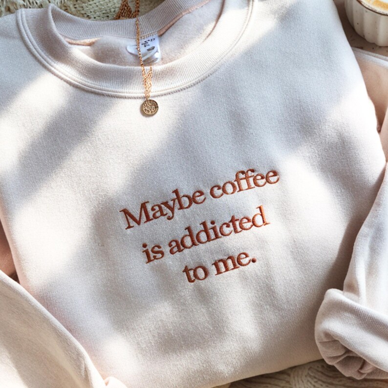May include: A light beige sweatshirt with the embroidered phrase "Maybe coffee is addicted to me." A gold necklace with a circular pendant is draped over the neckline. The sweatshirt has a crew neck and long sleeves.