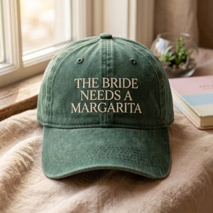 Puede incluir: Gorra de béisbol verde con el texto "THE BRIDE NEEDS A MARGARITA" en blanco. La gorra está hecha de una tela suave y texturizada y tiene una visera curva. La gorra está sobre una superficie beige.