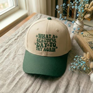 May include: A beige baseball cap with a green peak. The cap has the embroidered text "WHAT A BEAUTIFUL DAY TO TRY AGAIN" in green. The cap is displayed on a neutral-toned surface.