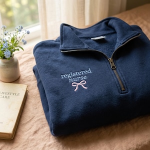 Könnte beinhalten: Ein marineblaues Sweatshirt mit Viertelreißverschluss und der gestickten Aufschrift "registered nurse" und einer rosa Schleife. Das Sweatshirt liegt gefaltet auf einem Bett mit einem Buch und einer kleinen Blumenvase im Hintergrund.