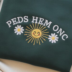 May include: Dark green sweatshirt with embroidered text "PEDS HEM ONC", a smiling yellow sun, and two white daisies. The design is centered on the front of the garment. The sweatshirt is made of a soft material.