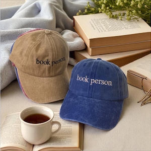 May include: Two baseball caps, one tan and one blue, embroidered with the words "book person". A cup of tea sits on an open book. The caps are on a table with other books.