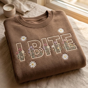 Könnte beinhalten: Ein braunes Sweatshirt mit dem Wort "BITE" in Creme gestickt, mit floralen Akzenten. Die Buchstaben sind mit bunten Blumen gefüllt und von gestickten Gänseblümchen umgeben. Das Sweatshirt hat einen Rundhalsausschnitt.