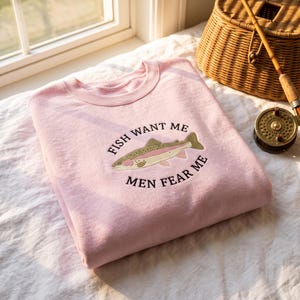 May include: A light pink crewneck sweatshirt featuring an embroidered graphic of a trout fish centered on the chest. The text "FISH WANT ME" is arched above the fish, and "MEN FEAR ME" is arched below the fish in black embroidered lettering.
