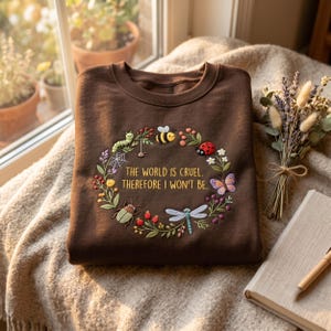 May include: A dark brown crewneck sweatshirt featuring an embroidered wreath of colourful insects, flowers, and a spiderweb. The text in the centre reads, "THE WORLD IS CRUEL. THEREFORE I WON'T BE." The design includes a bee, ladybug, and dragonfly.