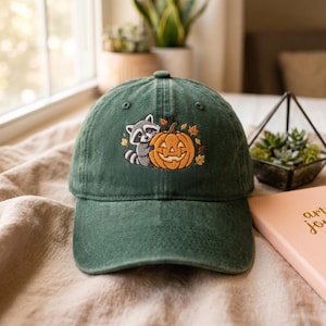 May include: A dark green baseball cap featuring an embroidered design of a grey and white raccoon hugging an orange jack-o'-lantern pumpkin, surrounded by small, scattered autumn leaves in shades of yellow, orange, and brown.