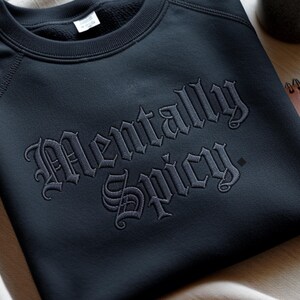 May include: A dark navy blue sweatshirt with the embroidered text "Mentally Spicy" in a gothic font. The sweatshirt has a crew neck and raglan sleeves. The fabric appears to be a soft, comfortable material.