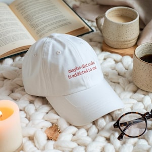 Peut inclure: Casquette de baseball blanche avec le texte brodé en rouge "maybe diet coke is addicted to me". La casquette est posée sur une surface blanche et texturée. D'autres éléments dans l'image incluent un livre, des tasses à café, une bougie et des lunettes.