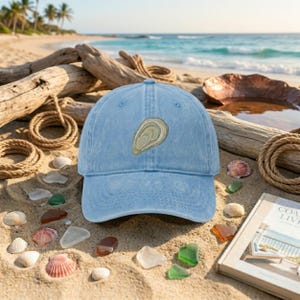 May include: A light blue baseball cap with a six-panel construction and a curved brim. The front center of the cap features an embroidered graphic of a single oyster shell in shades of beige, tan, and light green with detailed line work.