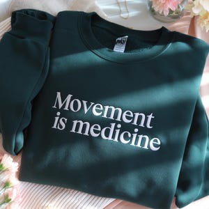 May include: A dark green sweatshirt with the phrase "Movement is medicine" in white lettering. The sweatshirt is folded, with the sleeve visible on the left side. The fabric appears soft and comfortable.