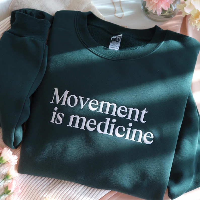 May include: A dark green sweatshirt with the phrase "Movement is medicine" in white lettering. The sweatshirt is folded, with the sleeve visible on the left side. The fabric appears soft and comfortable.