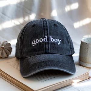 Könnte beinhalten: Eine schwarze Baseballkappe mit dem gestickten Text "good boy" in Weiß. Die Kappe liegt auf einem offenen Buch. Der Hut hat einen gebogenen Schirm und ein klassisches Sechs-Panel-Design.