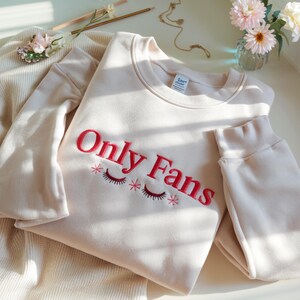 May include: A cream-colored sweatshirt with the embroidered text "Only Fans" in red, along with eyelash and star designs. The sweatshirt is folded and placed on a cream-colored surface.