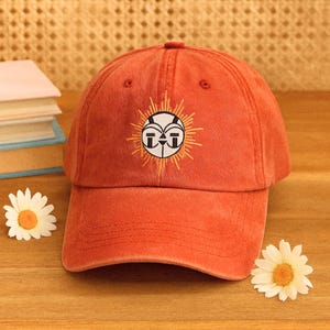 May include: A terracotta baseball cap with an embroidered sun design. The design features a white face with black details and yellow rays. The cap is on a wooden surface with a daisy. The cap is a fashion accessory.