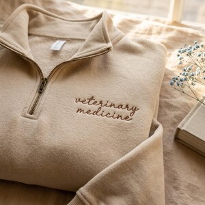Puede incluir: Sudadera beige con cremallera de cuarto de cremallera con el texto bordado "veterinary medicine" en marrón. La sudadera tiene cuello y puños acanalados, y una cremallera metálica. La prenda suave y de tonos neutros está doblada sobre una superficie clara.
