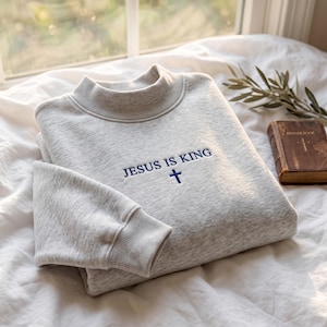 Könnte beinhalten: Ein hellgrau meliertes Langarm-Sweatshirt mit Stehkragen und gerippten Bündchen. Auf der Brust befindet sich ein dunkelblau gestickter Schriftzug JESUS IS KING, der direkt über einem kleinen, schlichten dunkelblauen Kreuz platziert ist.