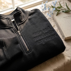 Peut inclure: Un sweat-shirt gris foncé avec une fermeture éclair quart de zip et l'inscription brodée "cute and medically expensive" sur le devant. Le sweat-shirt est plié, avec la fermeture éclair partiellement ouverte. Un livre et des fleurs sont en arrière-plan.