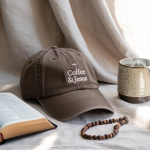 May include: A brown baseball cap embroidered with the words "Coffee & Jesus" and a cross. The image includes a mug, a rosary, and an open book, all arranged on a neutral-toned surface.