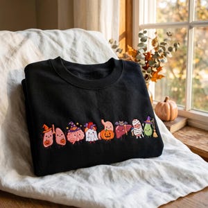 以下が含まれることがあります： ハロウィンの仮装をした人間の臓器の漫画風刺繍が施された黒いクルーネックのスウェットシャツ。肺、脳、心臓、胃、肝臓、アボカドが帽子をかぶったり、カボチャを持ったりしているデザインが中央に並んでいます。