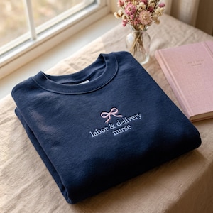 May include: Navy blue sweatshirt with a crew neck. The sweatshirt features a pink bow and the embroidered text "labor & delivery nurse". A pink notebook and a vase of dried flowers are in the background.