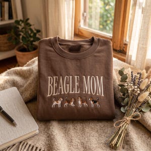 May include: A brown crewneck sweatshirt featuring the embroidered text BEAGLE MOM in a cream-coloured serif font, positioned above a row of four small, detailed embroidered beagle dogs in various standing and walking poses.