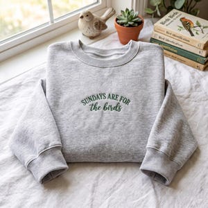 Op de afbeelding: Een lichtgrijs gemêleerd sweatshirt met ronde hals en donkergroene geborduurde tekst op de borst die "SUNDAYS ARE FOR the birds" leest in een combinatie van blokletters en cursief schrift, met lange mouwen en geribbelde boorden.