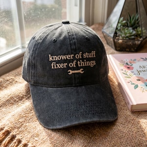 May include: A dark gray baseball cap with the embroidered text "knower of stuff fixer of things" and a wrench graphic. The cap is sitting on a textured surface near a window and a decorative terrarium.