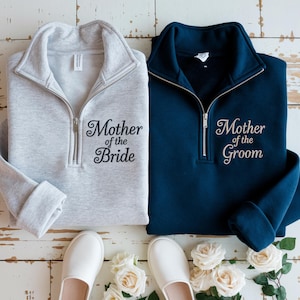 Pode incluir: Duas camisolas de quarto de fecho, uma cinza claro e outra azul marinho, bordadas com "Mother of the Bride" e "Mother of the Groom". Sapatos brancos e rosas dispostas por baixo.