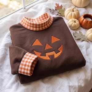 May include: A dark brown long-sleeve sweatshirt featuring an embroidered orange jack-o'-lantern face on the front. The garment has a mock neck collar and sleeve cuffs decorated with an orange and white gingham check pattern.